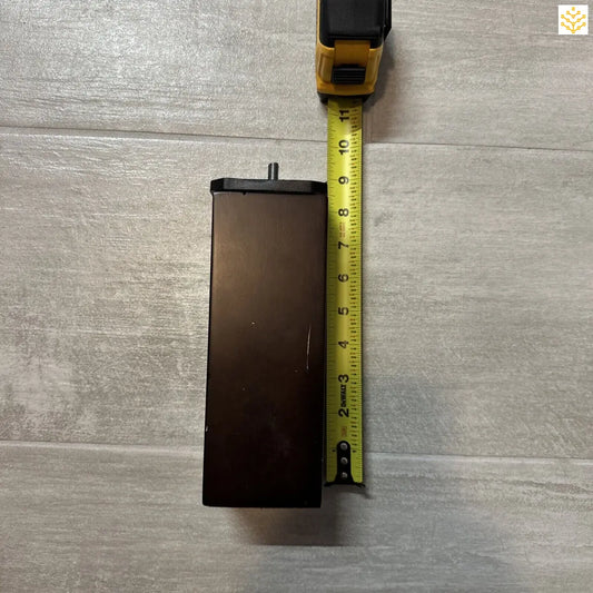 Rectangular, dark-colored electronic device or battery pack being measured with a yellow tape measure.